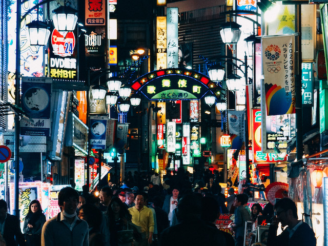 Enge Gasse voller Menschen von Kabukichō bei Nacht, bunte Neon-Lichter an den Häuserwänden links und rechts.