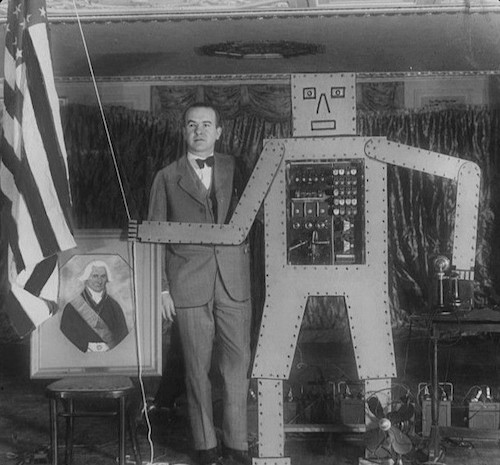 Schwarzweiß-Foto einer Tüftler-Werkstatt. Im Vordergrund ein Mann mit einem etwa 2 Meter großen Metallgestell in einfacher Menschenform, im Kern des Körpers ein Schaltkasten.