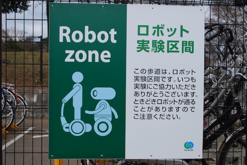 Hinweisschild auf Japanisch, dass in diesem Gebiet selbstständige Roboter unterwegs sein können. Dazu ein Bild mit zwei Robotern, die sich autonom bewegen.