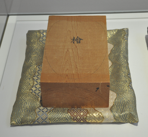 noch unbearbeiteter Holzblock, abgelegt auf einem wertvollen Kissen, auf dem Holzblock das Schriftzeichen für „hinoki“ „Zypresse“, mit Bleistift ein Oval als Maskenumriss aufgemalt