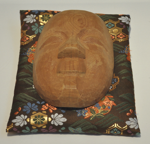 grob geschnitzte Nō-Maske, abgelegt auf einem wertvollen Kissen, Augen, Nasen, Mund erkennbar