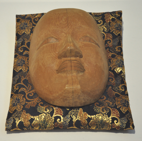 grob geschnitzte Nō-Maske, abgelegt auf einem wertvollen Kissen, Ausbuchtungen für Nase und Lippen fein herausgeschnitzt