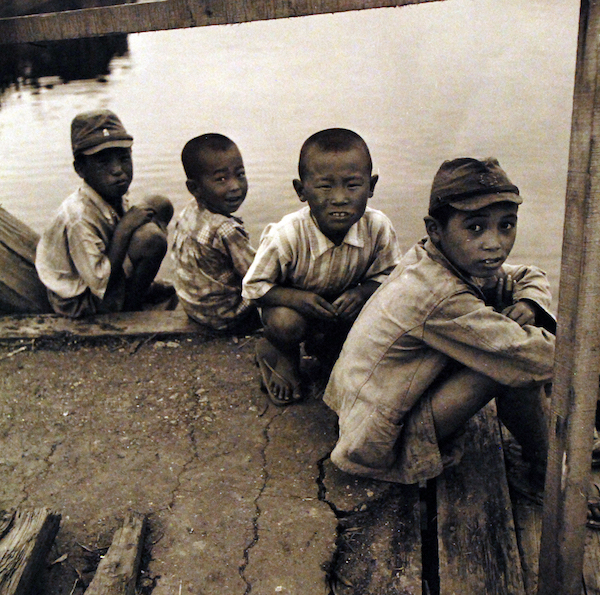 Auf einem Steg knien vier Jungs am Wasser und schauen neugierig in die Kamera