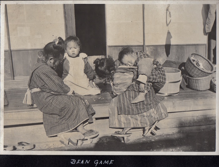 vier Kinder auf einer Holzveranda beim Spiel mit Bohnen als Spielsteinen, ein kleines Kind hat ein Baby auf dem Rücken