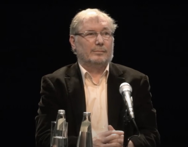Älterer Herr mit grauen Haaren und Vollbart vor schwarzem Hintergrund am Mikrofon eines Rednerpults.