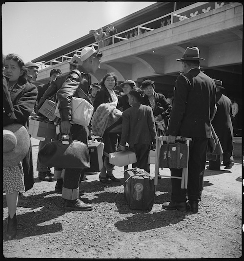 Japanerinnen und Japaner mit Koffern und Kisten in Warteschlange vor der Tribüne der Pferderennbahn.
