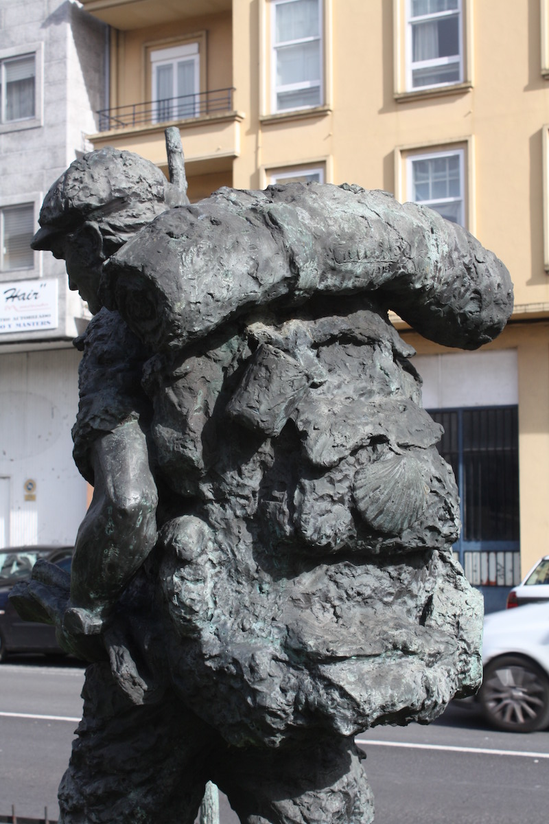 Bronzefigur eines gehenden Pilgers mit großem Rucksack, eingerollter Schlafmatte, Jakobsmuschel und Pilgerstab.