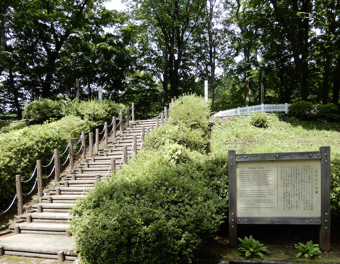 Treppen hinauf zu einem Grabhügel, an der Seite eine Tafel mit Erklärungen auf Englisch und Japanisch.