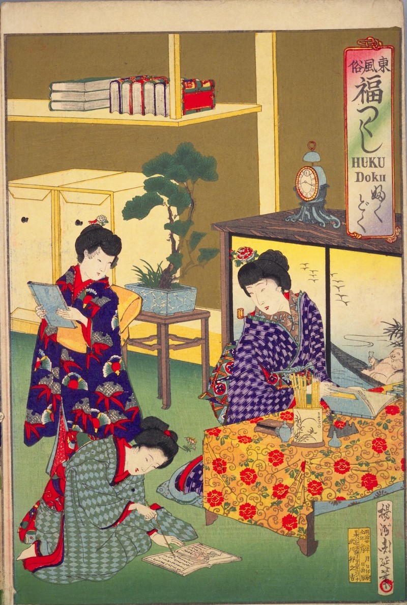 Drei junge Frauen: eine hockt am Boden und schreibt mit einem Pinsel, die beiden anderen schauen ihr zu. Im Hintergrund eine Regal mit Büchern.