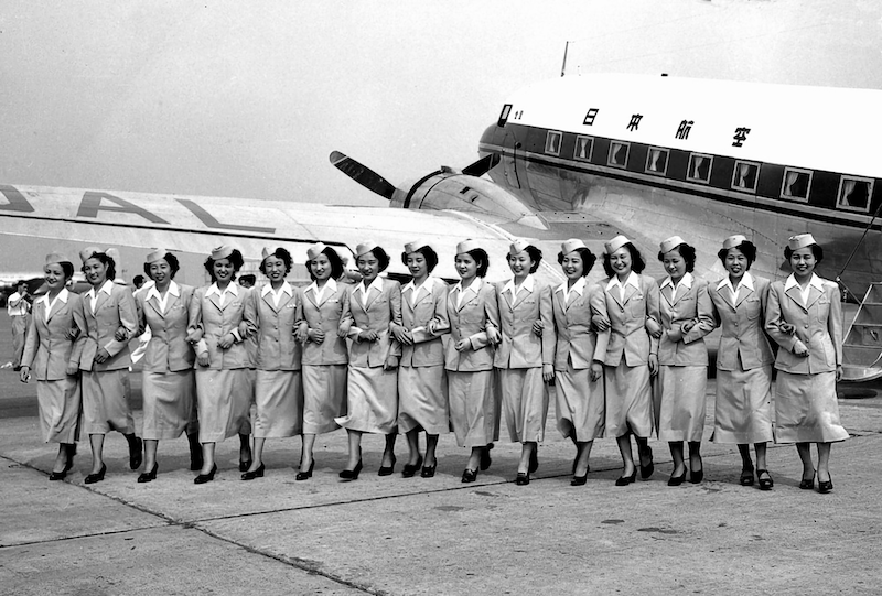 Schwarzweiß-Foto von 15 Flugbegleiterinnen, die sich untergehakt in einer Reihe vor einem Flugzeug posieren.
