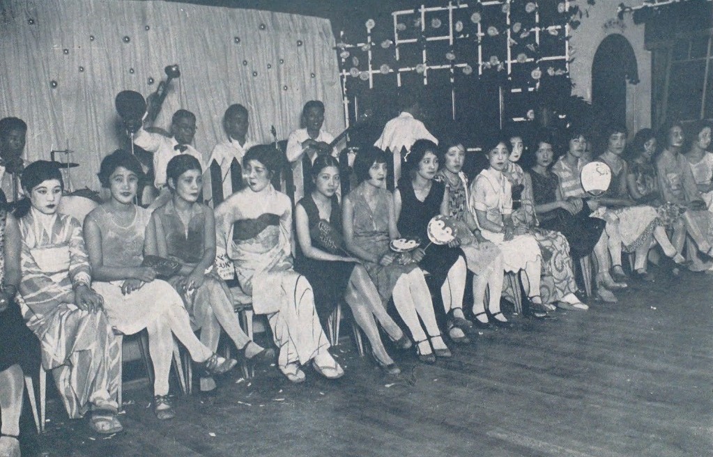 Moga: Schwarzweiß-Foto einer Tanzhalle, junge Frauen sitzen aufgereiht vor der Band