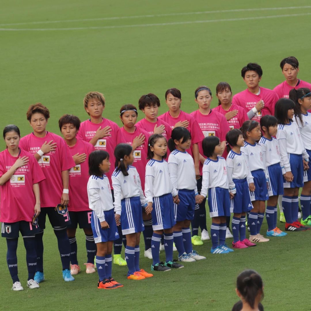 Die japanische Frauenmannschaft aufgereiht, vor ihnen Mädchen in Fußball-Trikots.
